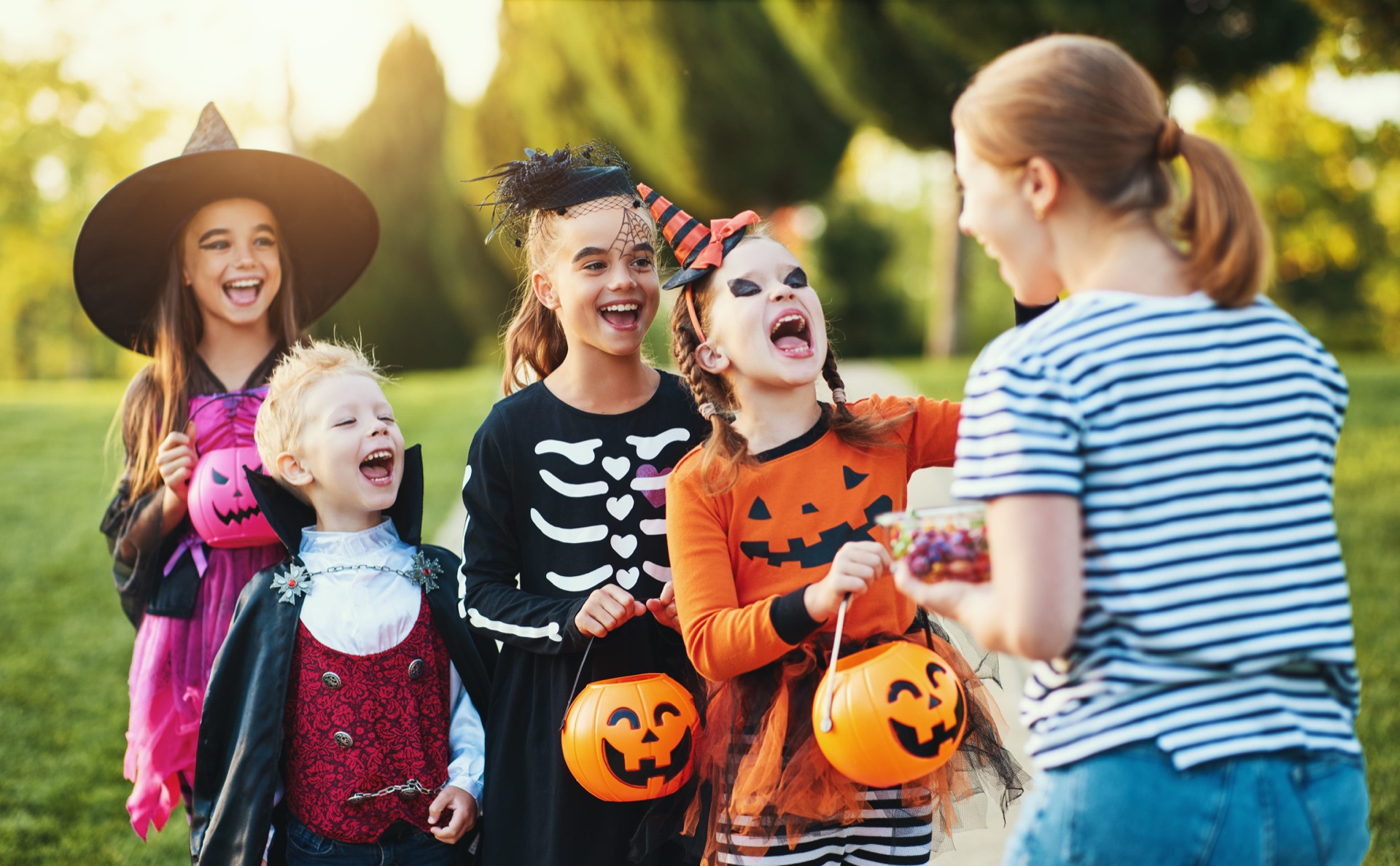 A group of laughing children in Halloween costumes holding jack-o-lantern candy buckets as an adult offers them candy