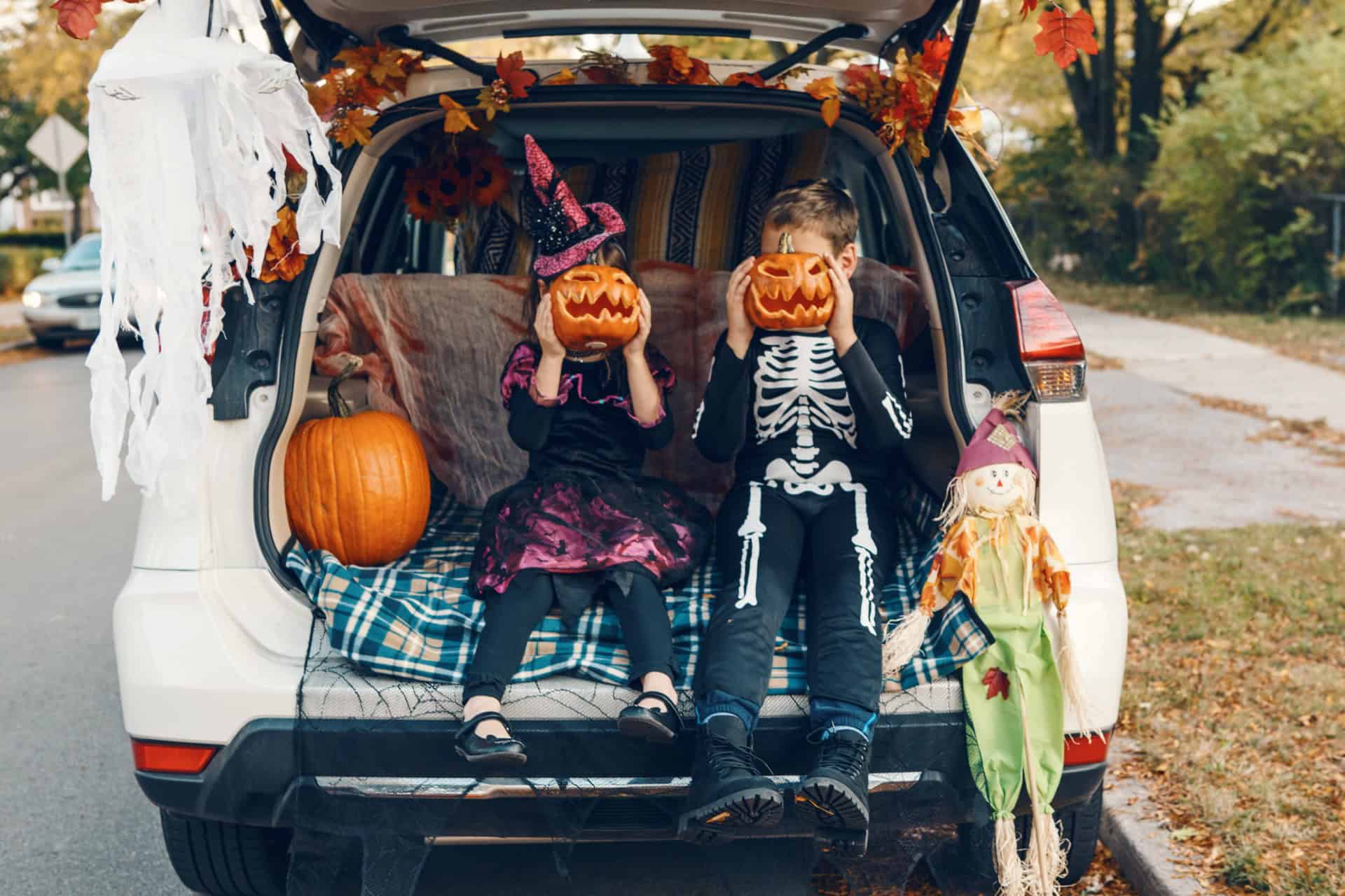 Two children in Halloween costumes sitting in a decorated car trunk holding carved jack-o-lanterns